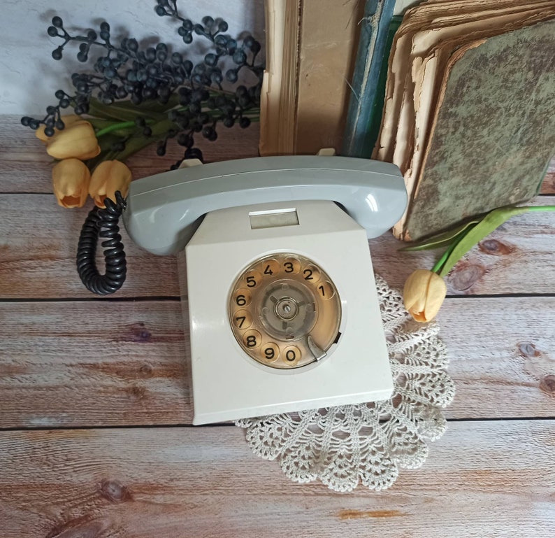 May include: A white rotary dial phone with a gray handset and a black cord. The phone has a white dial with black numbers 1 through 9 and 0. The phone is sitting on a wooden surface with a white doily.
