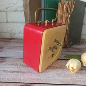 May include: A vintage red and cream-colored alarm clock with a gold-tone handle. The clock has a square shape with a cream-colored face and red sides. The clock is set against a backdrop of old books and a wooden surface.
