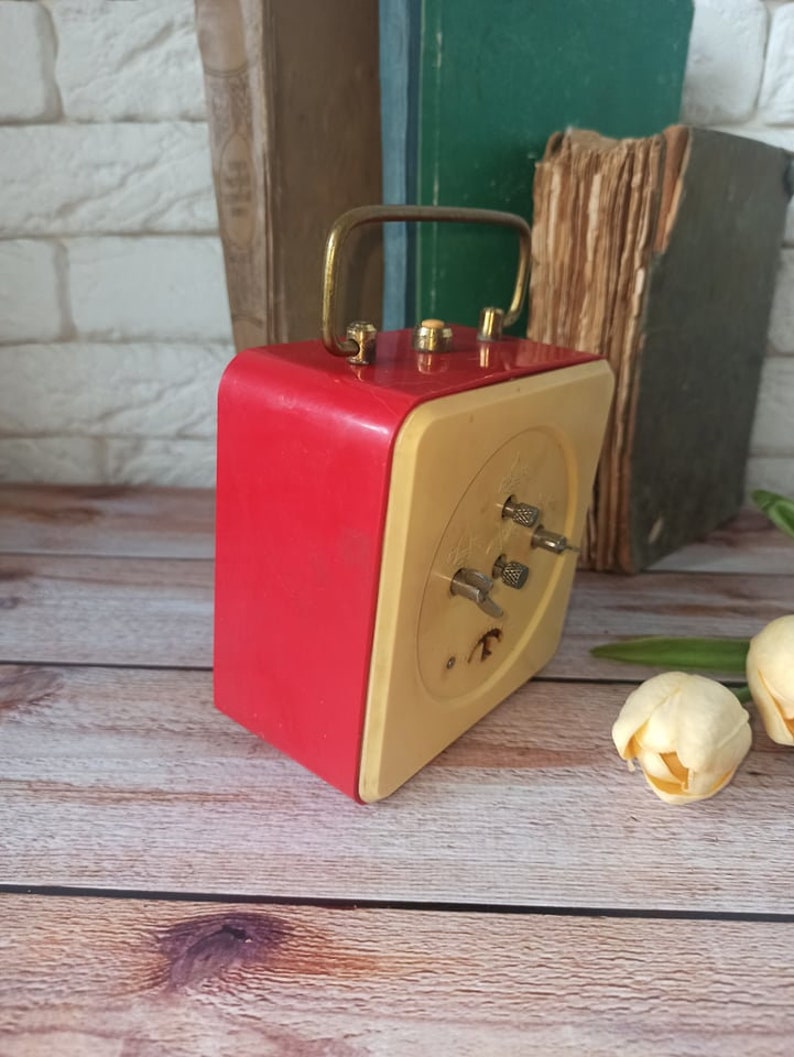 May include: A vintage red and cream-colored alarm clock with a gold-tone handle. The clock has a square shape with a cream-colored face and red sides. The clock is set against a backdrop of old books and a wooden surface.