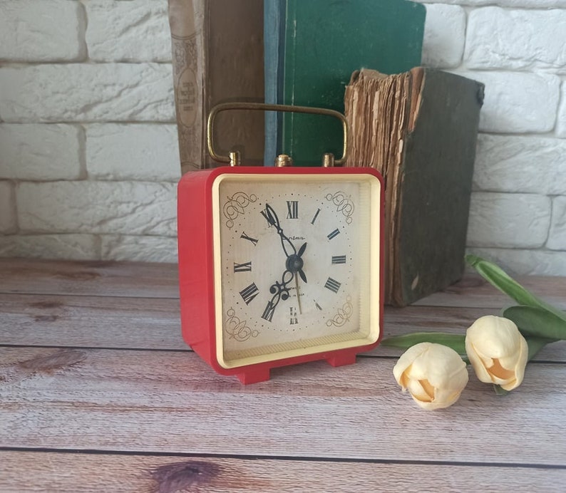 May include: A vintage red square alarm clock with a gold-colored handle and trim. The clock face has Roman numerals and decorative flourishes. The clock is set on a wooden surface, with books in the background.