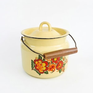 Puede incluir: Un jarro de leche de esmalte color crema con tapa y asa de madera. El jarro está decorado con un diseño floral en rojo, naranja y amarillo. El asa es marrón y el borde es negro.