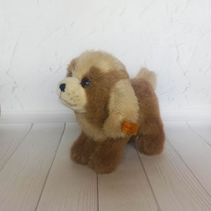 Puede incluir: Un perro de peluche marrón y crema con un botón Steiff en la oreja. El perro está de pie sobre una superficie de madera blanca.