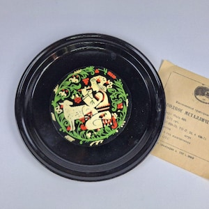 May include: A black, round decorative tray with a painted central design featuring a figure playing a flute next to a sheep, surrounded by green foliage and red accents. A paper with text is next to the tray.