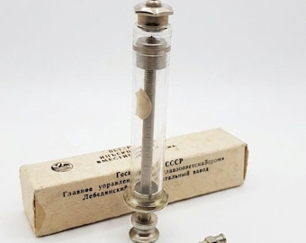 Puede incluir: Una jeringa de vidrio vintage con componentes metálicos, acompañada de una pequeña caja y varias agujas. La jeringa tiene un cilindro transparente con marcas de medición y un émbolo. La caja tiene texto cirílico.