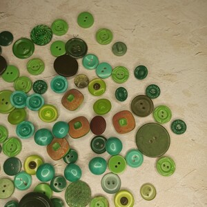 May include: A collection of various green buttons in different shades and sizes. The buttons include round, square, and textured designs, with colors ranging from light lime to deep forest green. Some buttons have two or four holes, while others have decorative patterns.