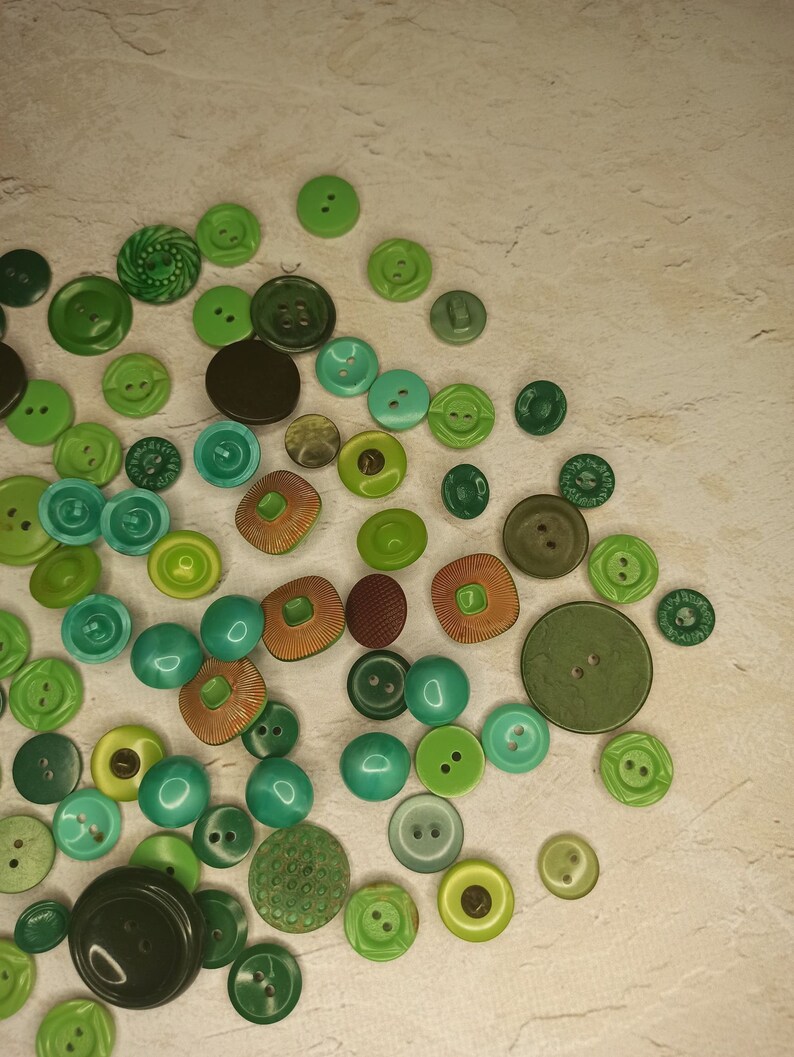 May include: A collection of various green buttons in different shades and sizes. The buttons include round, square, and textured designs, with colors ranging from light lime to deep forest green. Some buttons have two or four holes, while others have decorative patterns.