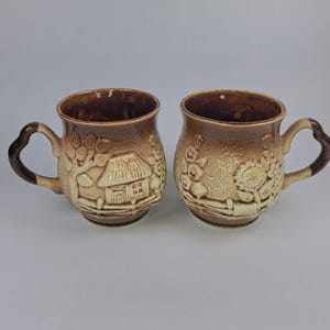 Puede incluir: Dos tazas de cerámica marrón con diseños en relieve. Una taza presenta una escena de casa, la otra un patrón floral. Ambas tazas tienen asas e interiores marrones oscuros, adecuadas para bebidas calientes.