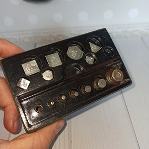 Puede incluir: Una caja de pl&aacute;stico negro con un juego de pesas de plata para una balanza. Las pesas son de diferentes formas y tama&ntilde;os, y est&aacute;n etiquetadas con su peso en gramos.