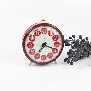 Op de afbeelding: Een vintage rode en crèmekleurige Lantas wekker met een ronde wijzerplaat. De klok heeft rode cijfers en wijzers, tegen een crèmekleurige achtergrond. De klok wordt ondersteund door drie kleine metalen pootjes.