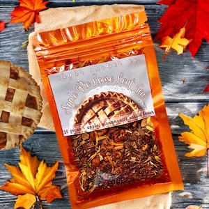 Könnte beinhalten: Ein oranger Beutel mit "Apple Pie Loose Leaf Tea" mit einem Tortengrafik, 20 Gramm, Apfel-, Zimt- und Vanillegeschmack. Herbstblätter und ein Stück Kuchen umgeben die Teeverpackung und schaffen ein Herbstthema.