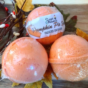 Pode incluir: Três bombas de banho laranja, embaladas em plástico transparente, são dispostas sobre uma superfície de madeira com decorações de outono. Uma bomba de banho tem um rótulo que diz "Sugar Shower Co. Pumpkin Pie Spice BATH BOMB!"