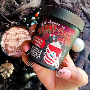 Pode incluir: Um recipiente preto de mistura para banho "Haunt Cocoa" com um gráfico vermelho e branco de uma bebida de chocolate quente. O rótulo inclui o texto "sugar shower co" e "hot chocolate scented fizzy bath mix". O recipiente é segurado em uma mão com nail art com tema de Halloween.