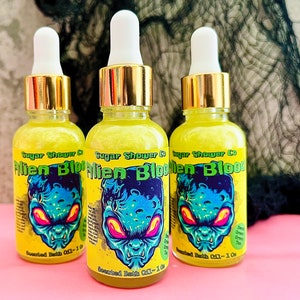 Könnte beinhalten: Drei kleine Glasflaschen mit goldenen Deckeln und gelber Flüssigkeit im Inneren. Die Flaschen sind mit "Sugar Shower Co Alien Blood Scented Bath Oil - 1 oz" beschriftet. Die Etiketten zeigen einen Comic-Alien mit grüner Haut und roten Augen.