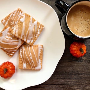 May include: Four square pastries with a white icing drizzle on a white plate. The pastries are next to a cup of coffee and two small orange pumpkins on a dark wood surface.