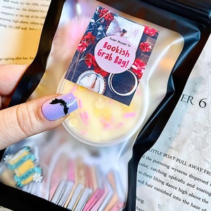 Peut inclure: Un sac en plastique transparent contenant un récipient rond avec de la cire jaune et des vermicelles roses, étiqueté "Bookish Grab Bag!". Le sac contient également des embellissements sur le thème des livres. L'arrière-plan comprend du texte d'un livre.