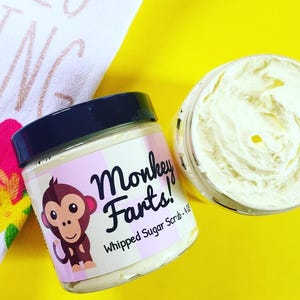 May include: Two jars of whipped sugar scrub on a yellow surface. One jar is open, revealing the creamy contents. The other jar has a black lid and a label with a cartoon monkey and the text "Monkey Farts!" The jar is next to a floral towel.