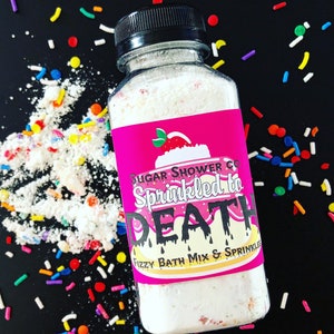 May include: A clear plastic bottle with a black cap, filled with white bath mix and sprinkles. The label is pink with the text "Sugar Shower Co. Sprinkled to DEATH Fizzy Bath Mix & Sprinkles". Colorful sprinkles are scattered around the bottle on a black surface.