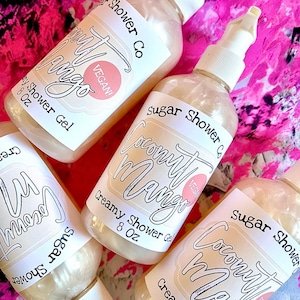 May include: Four clear plastic bottles of Sugar Shower Co. Coconut Mango Creamy Shower Gel, each containing 8 oz. The labels feature the product name in a script font, with a pink "VEGAN!" circle. The bottles are arranged on a pink and white patterned surface.