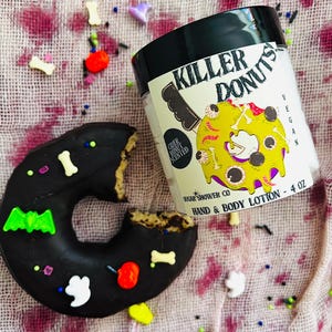 May include: A black and yellow hand and body lotion bottle with a spooky donut design. The label reads "Killer Donuts" and "Cider Donut Scented". The bottle is 4 oz and is vegan.
