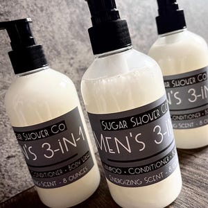 Puede incluir: Tres botellas de plástico transparente con dispensadores de bomba negros. Cada botella tiene una etiqueta gris con texto blanco que dice "Sugar Shower Co Men's 3-en-1 Champú, Acondicionador, Gel de ducha, Aroma energizante - 237 ml."
