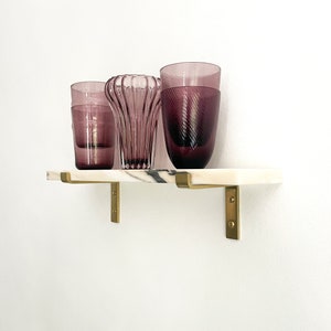 May include: Three glass vases in shades of purple, sitting on a white marble shelf with gold metal brackets.