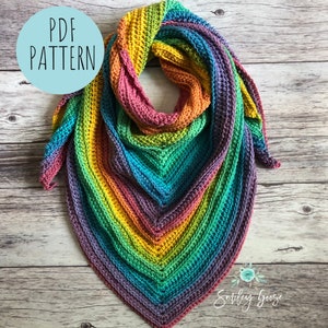 May include: A rainbow colored crochet shawl with a PDF pattern label. The shawl is made with a variety of colors, including red, orange, yellow, green, blue, and purple. The shawl is folded in half and laid out on a wooden surface.