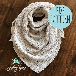 Puede incluir: Un chal de ganchillo de color crema con un delicado patrón de encaje. El chal está doblado y se muestra sobre una superficie de madera. El texto "PDF PATTERN" está escrito en un círculo azul en la esquina superior derecha de la imagen.