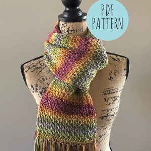 Op de afbeelding: Een gehaakte sjaal met franjes op een mannequin. De sjaal is in tinten groen, oranje en rood. De tekst "PDF PATTERN" staat op een blauwe cirkel in de rechterbovenhoek van de afbeelding.