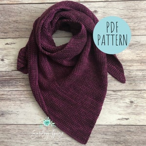 Puede incluir: Un chal triangular de punto de color burdeos con una etiqueta que dice "PDF pattern". El chal está colocado sobre una superficie de madera.