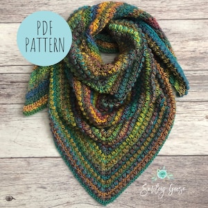 May include: A multi-coloured crochet shawl with a teal, green, brown and orange colour scheme. The shawl is folded and displayed on a wooden background. The text "PDF PATTERN" is on a blue circle in the top left corner of the image. The text "Smiling Goose" is in the bottom right corner of the image.