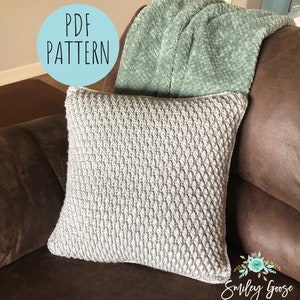 May include: A grey crocheted cushion with a textured pattern sits on a brown leather sofa. A light green throw blanket is draped over the back of the sofa. A blue circle with the text "PDF PATTERN" is in the top left corner of the image. The text "Smiley Goose HANDMADE" is in the bottom right corner of the image.