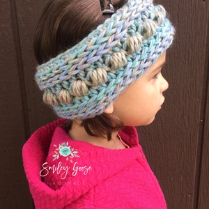 Puede incluir: Una diadema de punto azul y beige con pompones. La diadema la lleva puesta un ni&ntilde;o con el pelo casta&ntilde;o. El texto "Smiley Goose Handmade" es visible en la chaqueta rosa del ni&ntilde;o.