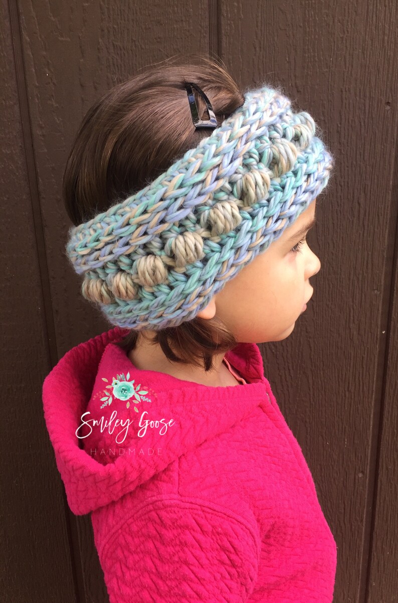 Puede incluir: Una diadema de punto azul y beige con pompones. La diadema la lleva puesta un ni&ntilde;o con el pelo casta&ntilde;o. El texto "Smiley Goose Handmade" es visible en la chaqueta rosa del ni&ntilde;o.