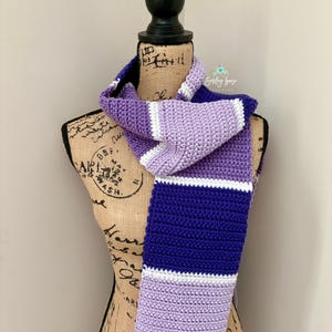 Puede incluir: Una bufanda tejida a ganchillo a mano con rayas horizontales en tonos morado oscuro, lavanda claro y blanco. La bufanda est&aacute; colocada sobre el torso de un maniqu&iacute;. Un peque&ntilde;o logotipo con el texto Smiling Gaze Handmade es visible arriba.