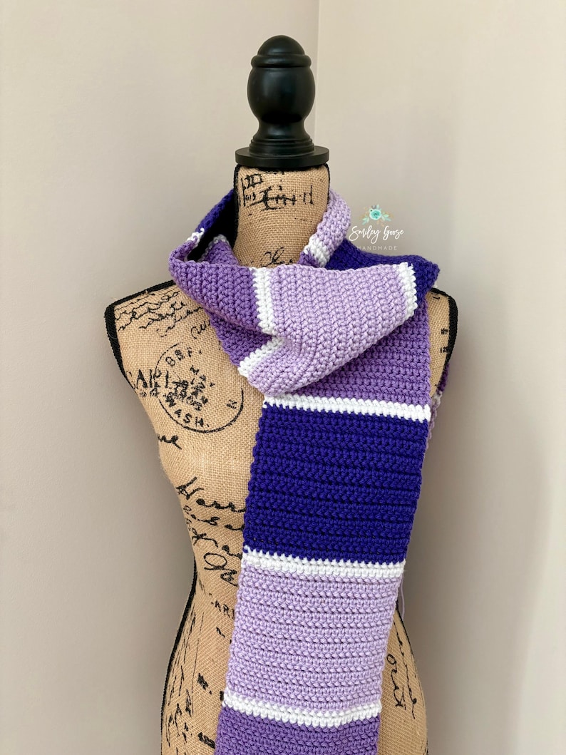 Puede incluir: Una bufanda tejida a ganchillo a mano con rayas horizontales en tonos morado oscuro, lavanda claro y blanco. La bufanda est&aacute; colocada sobre el torso de un maniqu&iacute;. Un peque&ntilde;o logotipo con el texto Smiling Gaze Handmade es visible arriba.