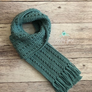 May include: A teal crocheted scarf with fringe on the ends. The scarf is laid out on a wooden surface.
