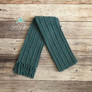 May include: A teal crocheted scarf with a fringe on one end. The scarf is laid out on a wooden surface.