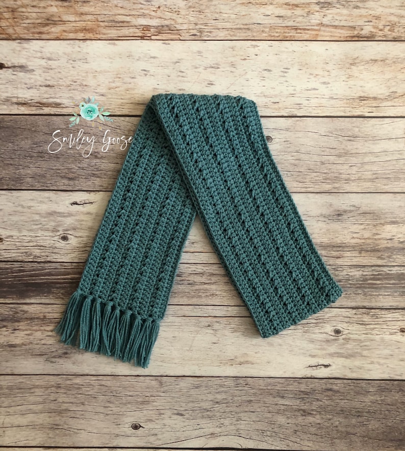 May include: A teal crocheted scarf with a fringe on one end. The scarf is laid out on a wooden surface.