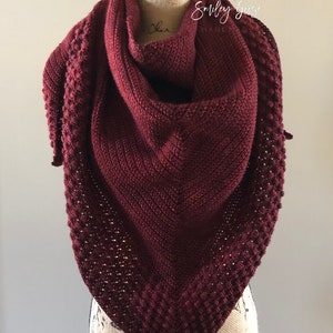 May include: A burgundy crocheted shawl with a textured pattern, displayed on a mannequin. The shawl is triangular in shape and has a delicate, lacy edge.