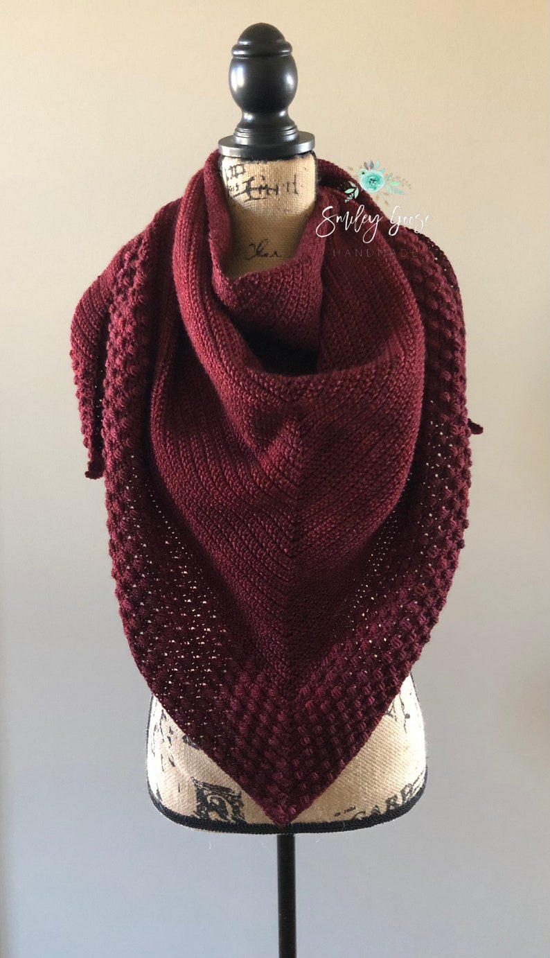May include: A burgundy crocheted shawl with a textured pattern, displayed on a mannequin. The shawl is triangular in shape and has a delicate, lacy edge.