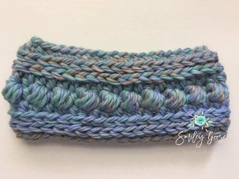 Puede incluir: Una diadema de crochet azul, verde y marr&oacute;n con un dise&ntilde;o texturizado. La diadema est&aacute; hecha por Smiley Goose Handmade.