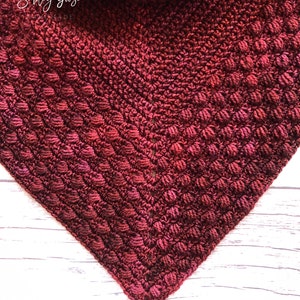 May include: Close-up of a crocheted triangle shawl in a deep red color with a gradient of lighter red hues. The shawl is made with a textured stitch that creates a bumpy surface.