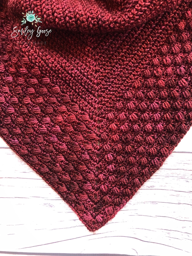 May include: Close-up of a crocheted triangle shawl in a deep red color with a gradient of lighter red hues. The shawl is made with a textured stitch that creates a bumpy surface.