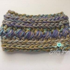 Puede incluir: Una diadema de crochet con un dise&ntilde;o texturizado en tonos de morado, verde y marr&oacute;n. La diadema est&aacute; hecha con un hilo grueso y tiene un borde trenzado.