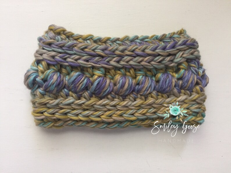 Puede incluir: Una diadema de crochet con un dise&ntilde;o texturizado en tonos de morado, verde y marr&oacute;n. La diadema est&aacute; hecha con un hilo grueso y tiene un borde trenzado.