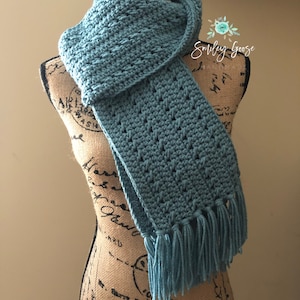 May include: A teal blue crocheted scarf with fringe on the ends. The scarf is draped over a fabric-covered mannequin.
