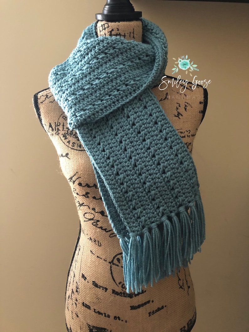 May include: A teal blue crocheted scarf with fringe on the ends. The scarf is draped over a fabric-covered mannequin.
