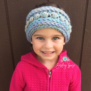 Puede incluir: Un ni&ntilde;o o ni&ntilde;a lleva una diadema de crochet azul, beige y blanca con una chaqueta rosa con capucha. La diadema tiene un dise&ntilde;o texturizado y abultado. El texto "Smiley Goose Handmade" es visible en la chaqueta.
