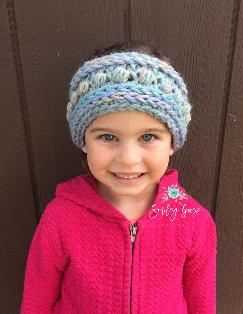 Puede incluir: Un ni&ntilde;o o ni&ntilde;a lleva una diadema de crochet azul, beige y blanca con una chaqueta rosa con capucha. La diadema tiene un dise&ntilde;o texturizado y abultado. El texto "Smiley Goose Handmade" es visible en la chaqueta.