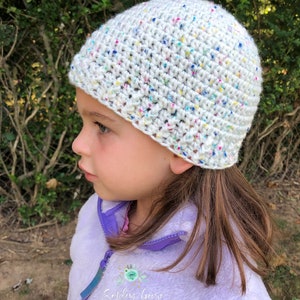 May include: A white crochet hat with colorful flecks. The hat is worn by a child with long brown hair. The child is wearing a purple fleece jacket with the text "Smiling Goose" embroidered on the front.